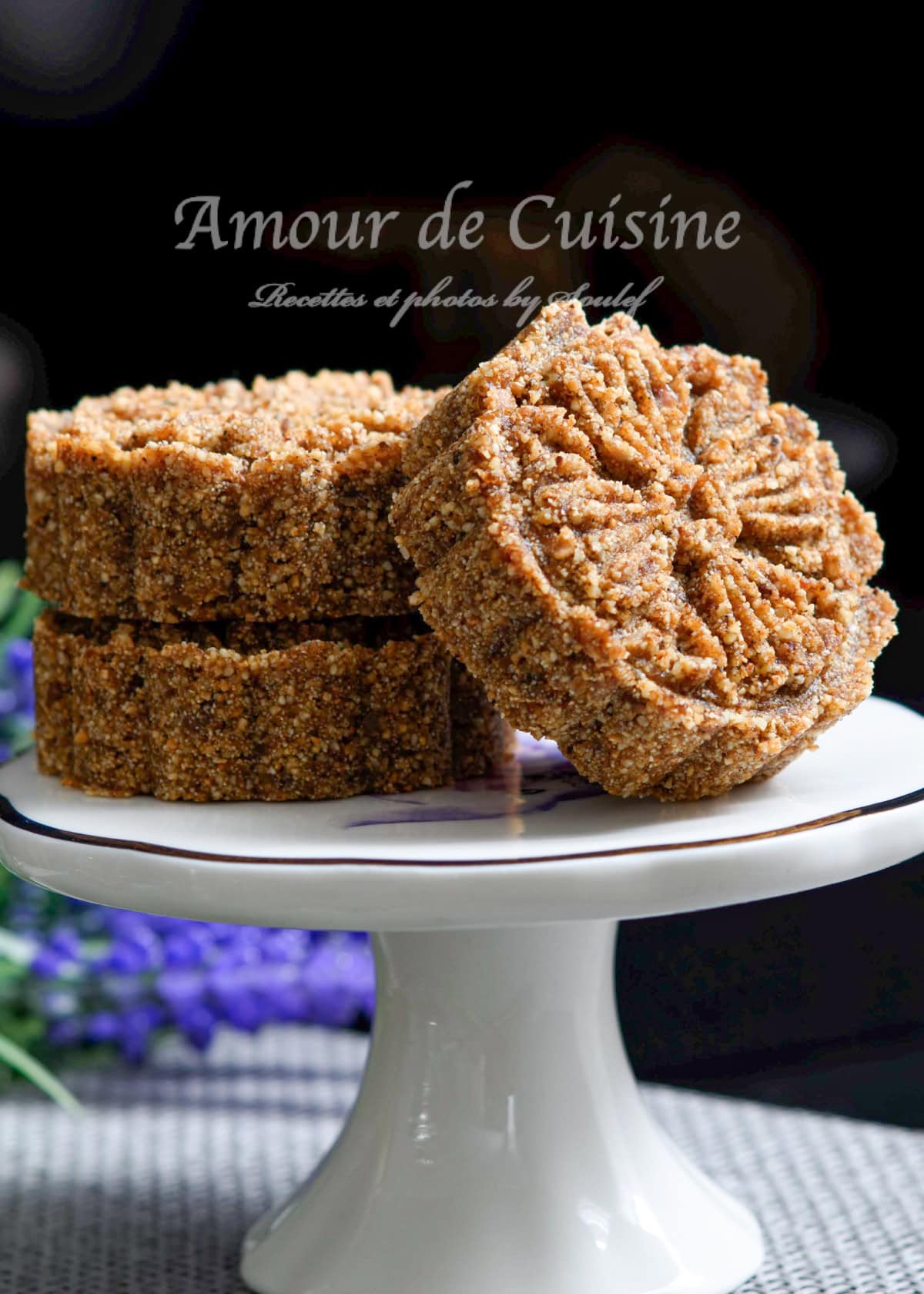 Rfiss tounsi, un gâteau algérien traditionnel aux dattes et à la semoule, présenté sur un présentoir blanc avec un décor floral en arrière-plan.