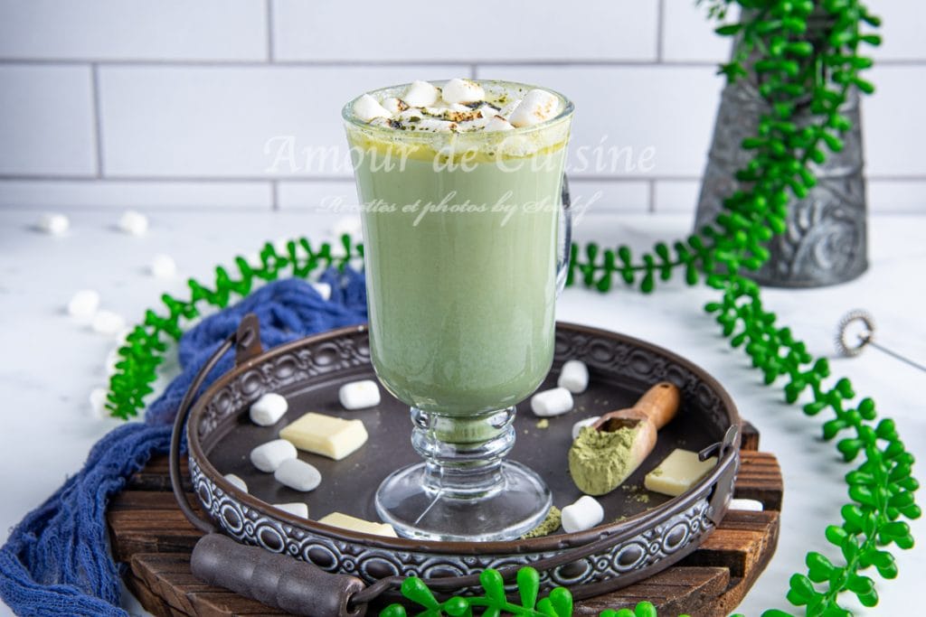 Verre de chocolat chaud au matcha et chocolat blanc sur un plateau en métal, entouré de morceaux de chocolat blanc, de marshmallows et de poudre de matcha